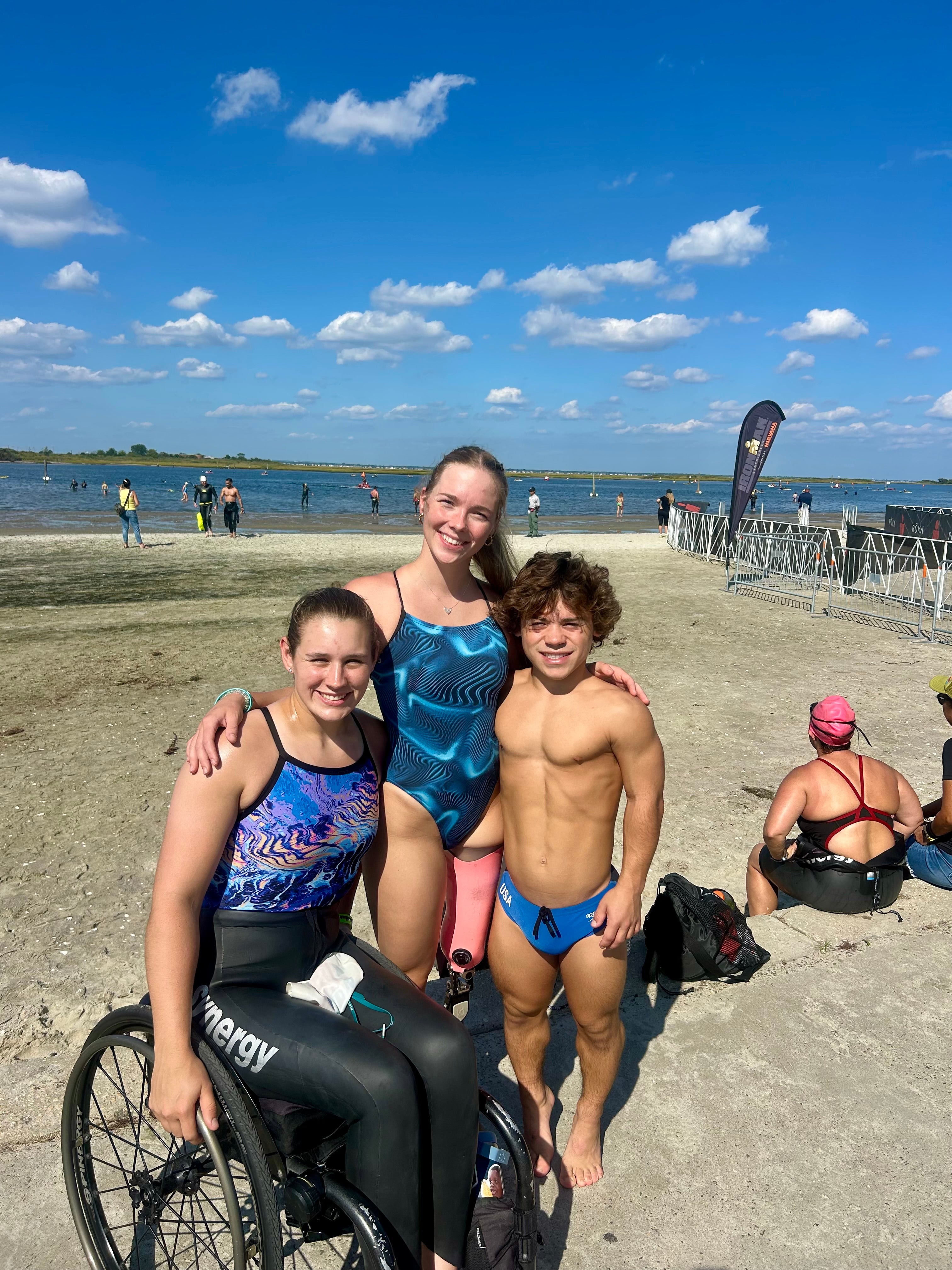 Three people, including one in a wheelchair, posing on a beach with a clear blue sky.

